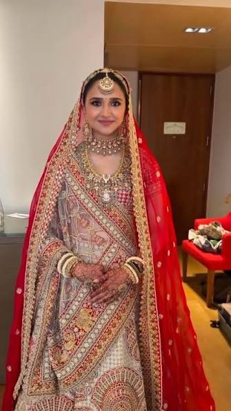 Dweta Patel standing in a red and gold bridal lehenga with an embroidered dupatta, maang tikka, layered gold and kundan necklaces and mehndi on both hands.