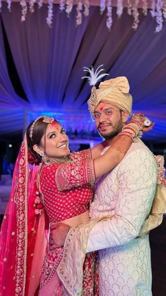 Dweta Patel and Karan Nathwani photographed together in wedding attire beneath a floral mandap, she in a red and pink lehenga and he in an ivory sherwani and safa.