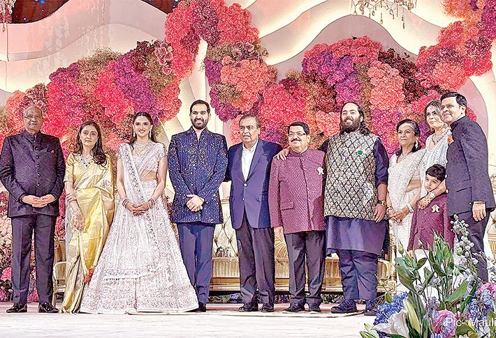 The wedding party and guests standing together on stage in formal Indian dress before a tall backdrop of pink, red and magenta flowers at the Nathwani wedding reception in Mumbai.