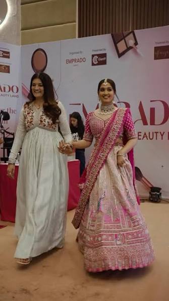 Two women walking a showcase floor at a bridal and beauty exhibition, one in a pink and ivory embroidered bridal lehenga with a draped dupatta and layered jewellery, the other in a cream and red embroidered outfit.